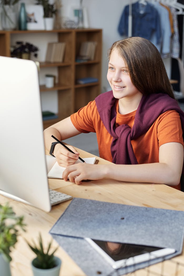 Services Smiling teen girl studying online at her home desk with a computer and notepad.