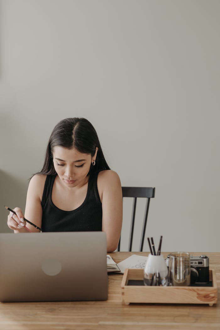 Services Asian woman at home desk engaged in online learning with laptop, notes, and pen.